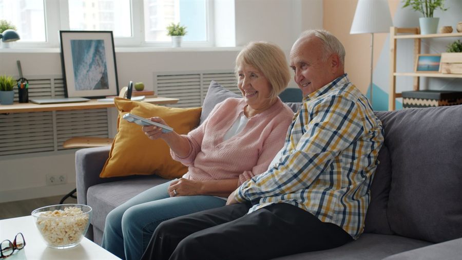 Older couple on a sofa with popcorn, one holding the TV remote